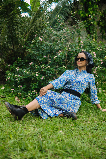 une femme asiatique dans une robe bleue et des lunettes de soleil est assis sur l'herbe verte pleine de fleurs le matin - Photo, image