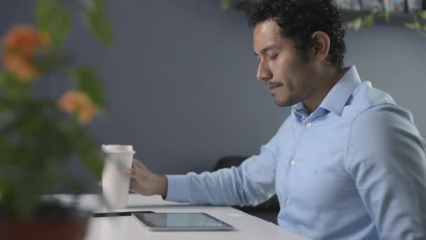 Νεαρός ενήλικας με καφέ δέρμα και σγουρά μαλλιά κάνει το γραφείο στο σπίτι στο tablet και καφέ του, στη δουλειά του - Πλάνα, βίντεο