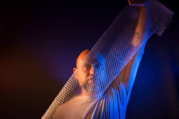 Bald bearded man stretching a bubble wrap in front of his face. Isolated on dark red background. - Photo, Image