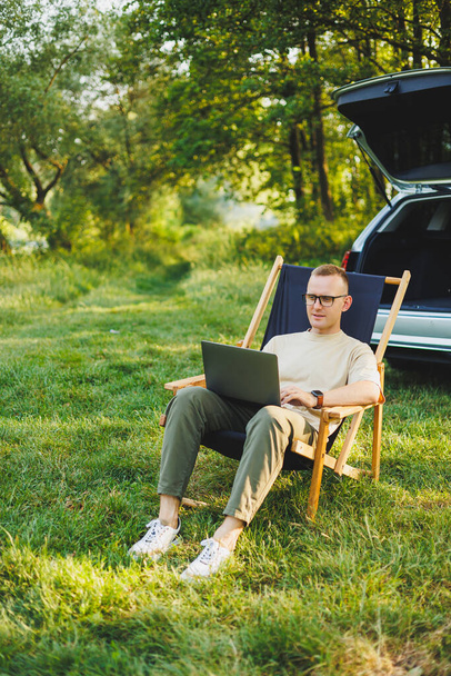 Веселый человек сидит на стуле в природе и работает онлайн на ноутбуке. Человек путешествует и работает дистанционно на ноутбуке. Работа в офисе на природе - Фото, изображение