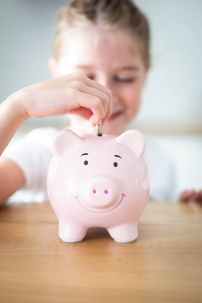 Καυκάσιος κορίτσι βάζει ένα νόμισμα σε ένα ροζ κουμπαρά. Εξοικονόμηση χρημάτων για παιδιά. Κάθετη βολή. - Φωτογραφία, εικόνα