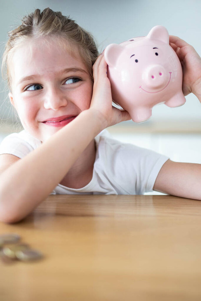 Gelukkig blank kind meisje schudden een glimlachende roze spaarvarken bank in haar handen op een houten tafel. Besparen geld voor kinderen concept. Verticaal schot. - Foto, afbeelding