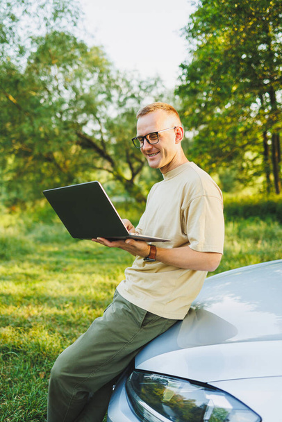 A cheerful man in glasses sits on the hood of his car in nature and works on a laptop. Freelance work, remote work during vacation - Foto, imagen