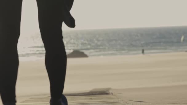 Un hombre con traje de neopreno camina por la playa sosteniendo una tabla de surf. Mid shot - Imágenes, Vídeo