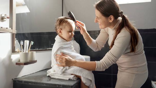 Bonne mère souriante peigner ses cheveux de bébé fils avec une brosse à cheveux après le lavage dans le bain. Concept de parentalité, bonheur et hygiène de l'enfant - Photo, image