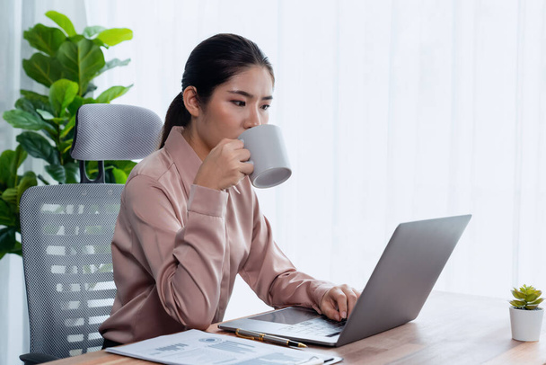 Młoda azjatycka, entuzjastyczna bizneswoman pracująca w nowoczesnym biurze, pijąca kawę, pilnie pracująca na laptopie, reprezentuje profesjonalnego i atrakcyjnego pracownika biurowego w swojej przestrzeni roboczej.. - Zdjęcie, obraz