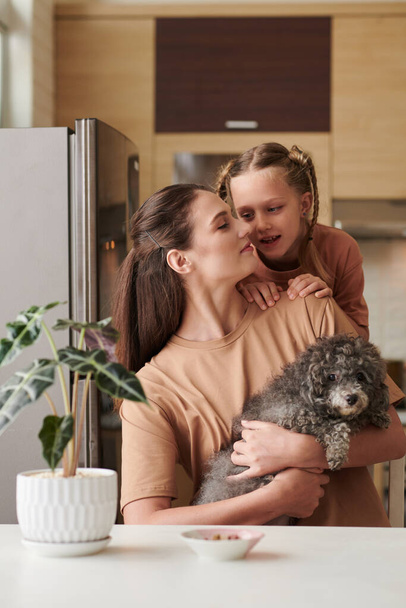 Girl telling something to mother sitting at kitchen table with small dog in hands - Photo, Image