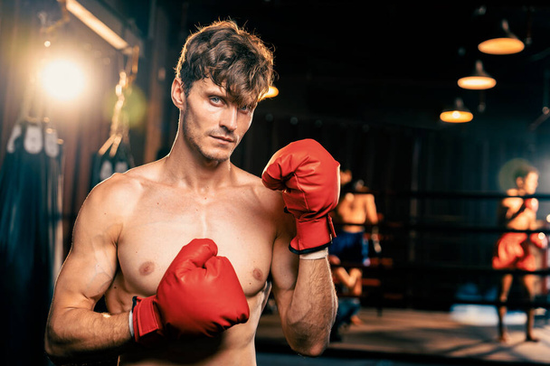 Боксер без рубашки позирует, кавказский боксер в красной перчатке в оборонительной стойке караула, готовый драться и бить в спортзале рингом и боксерским оборудованием на заднем плане. Импет - Фото, изображение