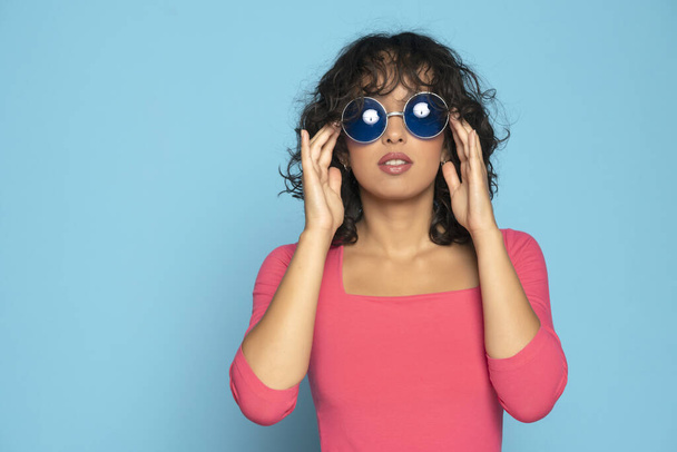 Young exotic brunette woman in pink blouse posing with a sunglasses on a blue studio background - Foto, immagini