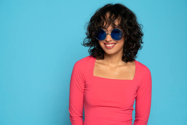 Pembe bluzlu, egzotik, gülümseyen esmer kadın mavi bir stüdyoda güneş gözlüğü takmış poz veriyor. - Fotoğraf, Görsel