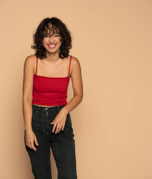 Молодая экзотическая счастливая брюнетка в красной рубашке без рукавов и джинсах позирует на фоне бежевой студии - Фото, изображение