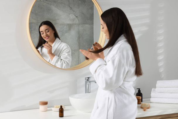 Happy young woman applying essential oil onto hair near mirror in bathroom - Фото, изображение