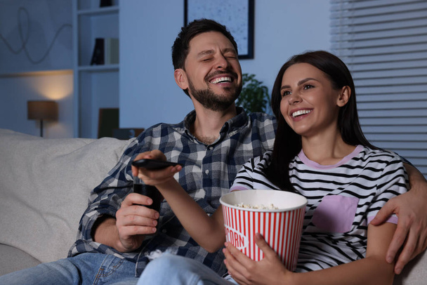 Happy couple watching show at home in evening. Woman holding popcorn and changing TV channels with remote control - Foto, Imagem