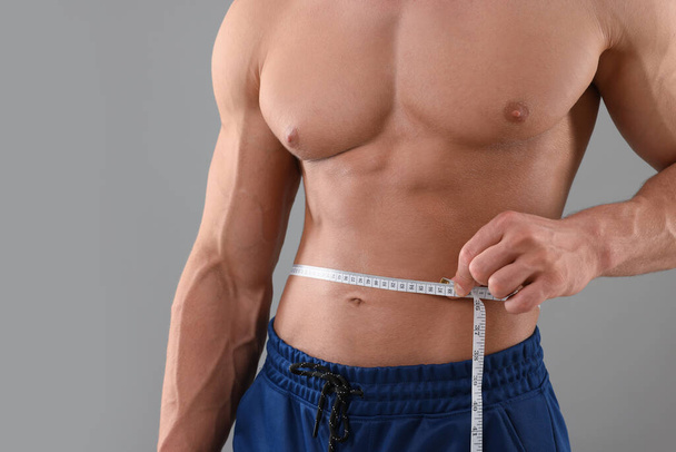 Athletic man measuring waist with tape on grey background, closeup. Weight loss concept - Foto, afbeelding