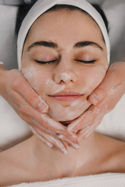 Bovenste weergave van een mooie jonge blanke vrouw leunend op een spa bed met gesloten ogen met een gezichtsmassage met schuim voor het verwijderen van dode huid. - Foto, afbeelding