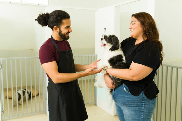 Femme latine parlant à un homme heureux travailleur apportant son chien shih tzu mignon à l'hôtel pour animaux de compagnie ou garderie pour un séjour  - Photo, image