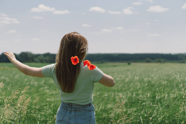 Жінка тримає червоні макові квіти на фоні поля. Атмосферний спокійний момент, сільське повільне життя. Жінка збирає квіти і відпочиває в літній сільській місцевості
 - Фото, зображення