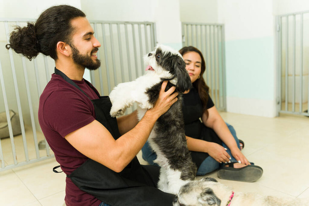 Joven latino abrazando y acariciando a un hermoso perro shih tzu mientras trabaja como peluquero de mascotas u hotel de guardería - Foto, Imagen