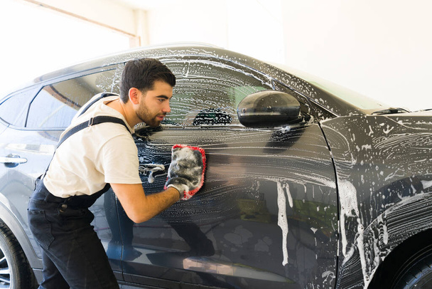 Atractivo joven latino lavando el coche de un cliente con un guante de lavado de autos y espuma en el lavado de autos - Foto, imagen