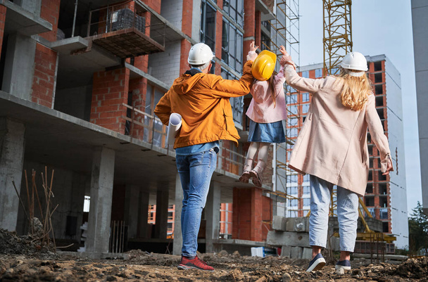 Back view of man and woman in safety helmets holding hands of adorable daughter and lifting kid in the air at construction site. Family with child standing outside building under construction. - 写真・画像