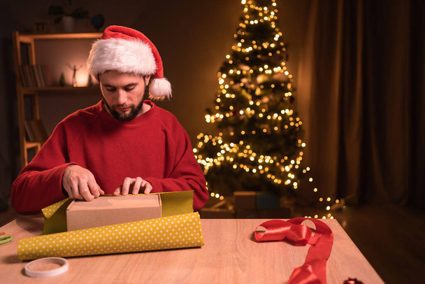 Szakállas férfi karácsonyi ajándékokat csomagol papírba, miközben otthon ül az asztalnál esténként. Fénymásolási hely - Fotó, kép