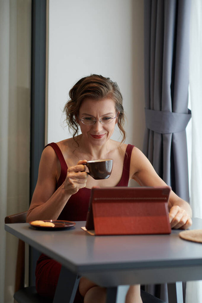 Χαμογελώντας elelelegant ώριμη γυναίκα πίνοντας πρωινό καφέ και διαβάζοντας ειδήσεις στον υπολογιστή tablet - Φωτογραφία, εικόνα