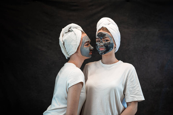 Girls friends in white towel applying black mud clay facial mask isolated on black. Anti age treatment, skincare - Photo, Image