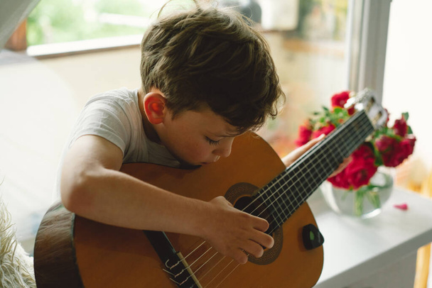 Cute boy uczy się grać na gitarze klasycznej na parapecie w pobliżu okna. Przytulny dom. Letnie wakacje styl życia. - Zdjęcie, obraz