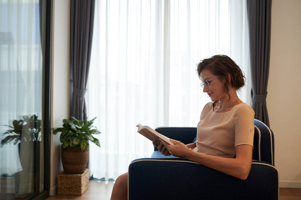 Положительная женщина средних лет читает книгу дома, когда отдыхает на диване - Фото, изображение