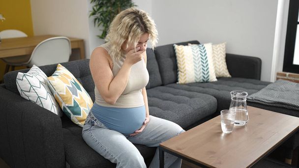 Молодая беременная женщина сидит на диване и страдает тошнотой дома - Фото, изображение