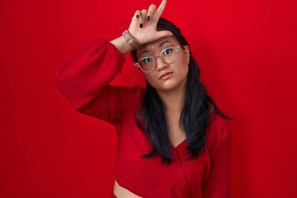 Aziatisch jong vrouw staande over rode achtergrond maken plezier van mensen met vingers op het voorhoofd doen verliezer gebaar spotten en beledigend.  - Foto, afbeelding