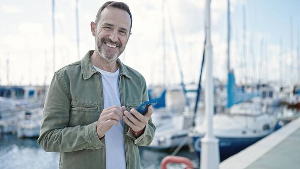 Мужчина средних лет уверенно улыбается, используя смартфон в порту - Фото, изображение