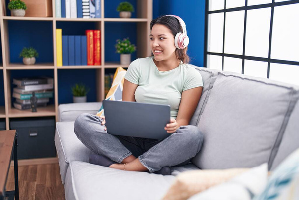 Latino nuori nainen käyttää kannettavaa tietokonetta kotona yllään kuulokkeet katselee pois puolelta hymyillen kasvot, luonnollinen ilme. nauraen luottavainen.  - Valokuva, kuva