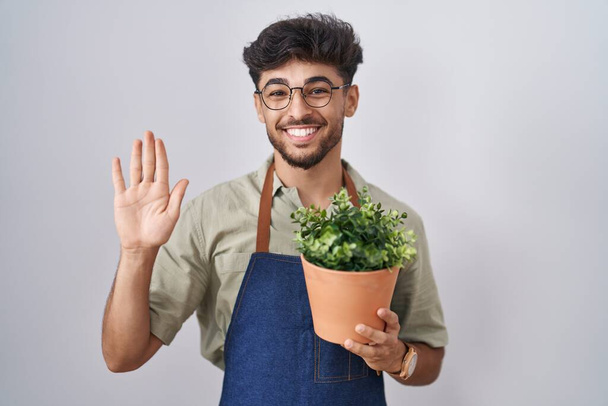Araber mit Bart hält grünen Pflanzenkübel in der Hand und verzichtet auf fröhliches Hallo und lächelnde freundliche Willkommensgeste  - Foto, Bild