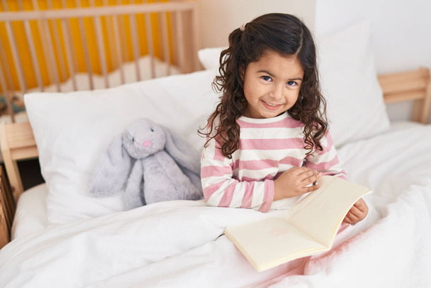 Adorable hispanique fille lecture livre assis sur le lit dans la chambre - Photo, image