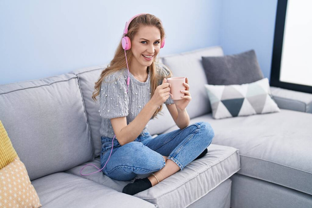 Mujer rubia joven escuchando música tomando café en casa - Foto, Imagen