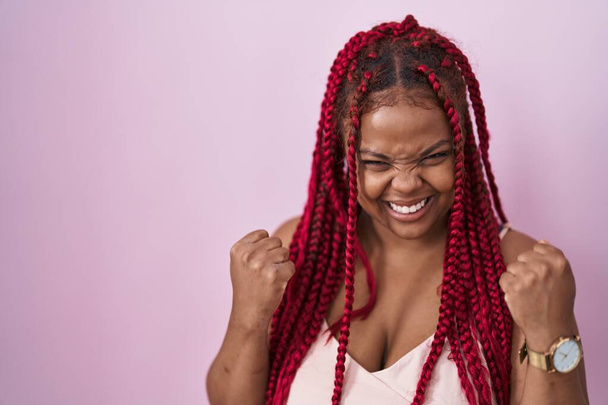 Femme afro-américaine avec les cheveux tressés debout sur fond rose très heureux et excité de faire geste gagnant avec les bras levés, souriant et criant pour le succès. concept de célébration.  - Photo, image