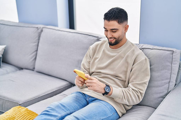 Young arab man using smartphone sitting on sofa at home - 写真・画像