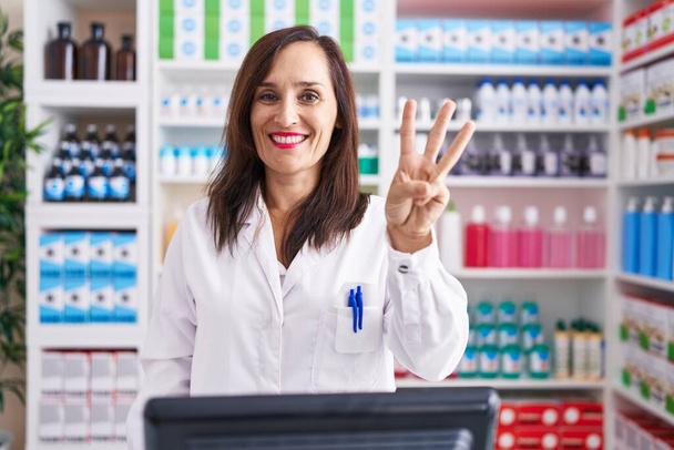 Middelbare leeftijd brunette vrouw werken bij apotheek drogisterij tonen en wijzen met vingers nummer drie, terwijl glimlachen zelfverzekerd en gelukkig.  - Foto, afbeelding