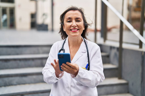 Middle age woman wearing doctor uniform using smartphone at street - Photo, Image