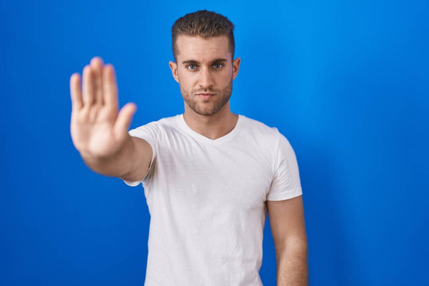 Jeune homme caucasien debout sur fond bleu faire arrêter chanter avec la paume de la main. expression d'avertissement avec geste négatif et sérieux sur le visage.  - Photo, image