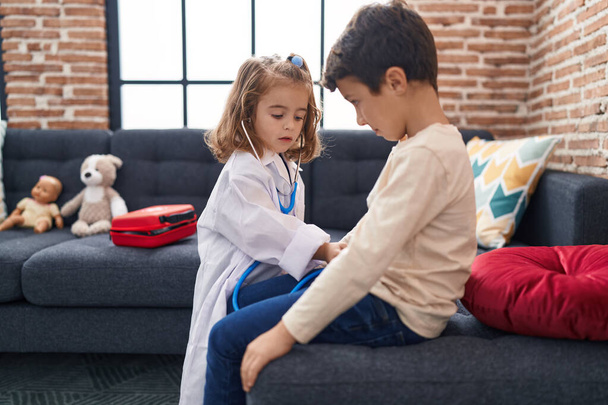 Imádnivaló fiú és lány orvosi egyenruhát visel, sztetoszkóppal vizsgálva otthon. - Fotó, kép