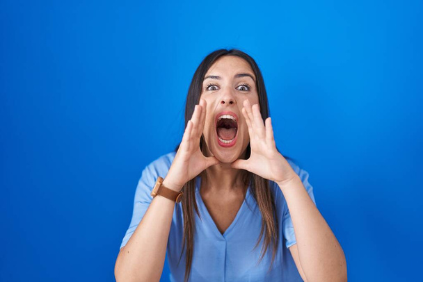 Junge brünette Frau steht vor blauem Hintergrund und schreit wütend laut mit den Händen über dem Mund  - Foto, Bild