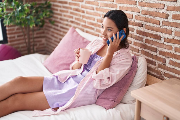 Junge hispanische Frau telefoniert mit Smartphone und trinkt Kaffee im Schlafzimmer - Foto, Bild