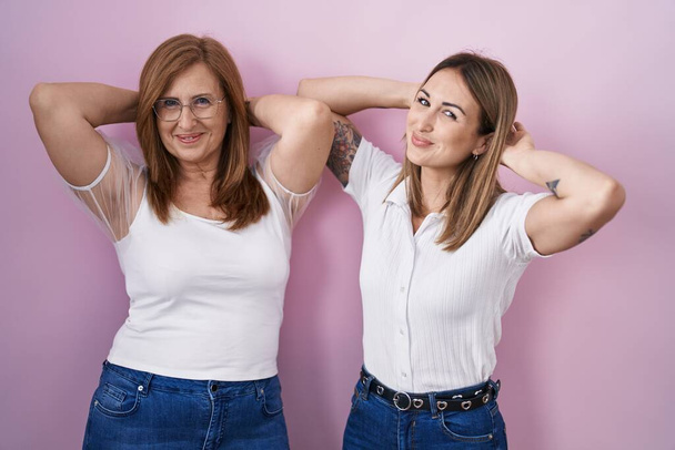 Ισπανίδα μητέρα και κόρη φορώντας casual λευκό t πουκάμισο πάνω από ροζ φόντο χαλάρωση και τέντωμα, τα χέρια και τα χέρια πίσω από το κεφάλι και το λαιμό χαμογελώντας χαρούμενος  - Φωτογραφία, εικόνα