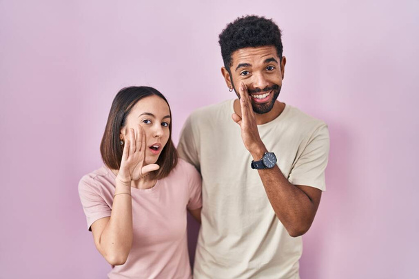 Junge hispanische Paar zusammen über rosa Hintergrund Hand auf Mund erzählen geheime Gerüchte, flüstern böswillige Gespräch  - Foto, Bild