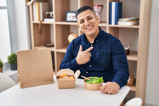 Joven hispano comiendo llevar comida sonriendo feliz señalando con la mano y el dedo  - Foto, imagen