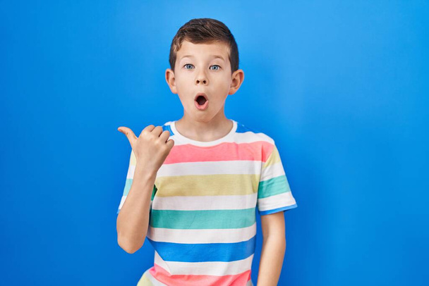 Young caucasian kid standing over blue background surprised pointing with hand finger to the side, open mouth amazed expression.  - Photo, Image