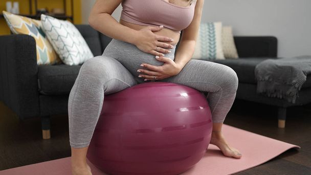 Nuori raskaana oleva nainen tekee valmistautumisharjoituksen istuen hyvässä kunnossa pallolla kotona - Valokuva, kuva
