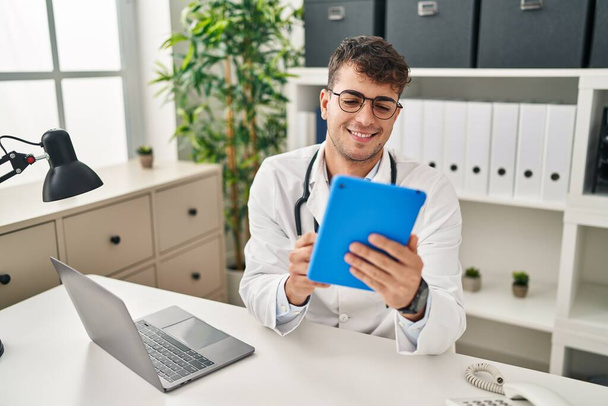 Jonge Spaanse man arts met behulp van laptop en touchpad werken in de kliniek - Foto, afbeelding